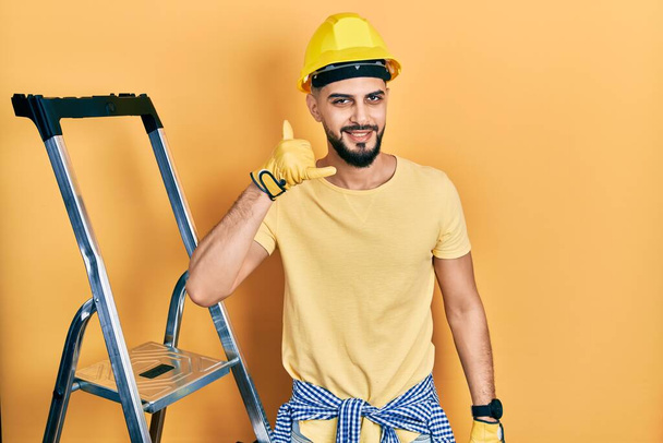 Красавчик с бородой по строительной лестнице в каске, улыбаясь, делает телефонный жест рукой и пальцами, как будто разговаривает по телефону. коммуникационные концепции.  - Фото, изображение