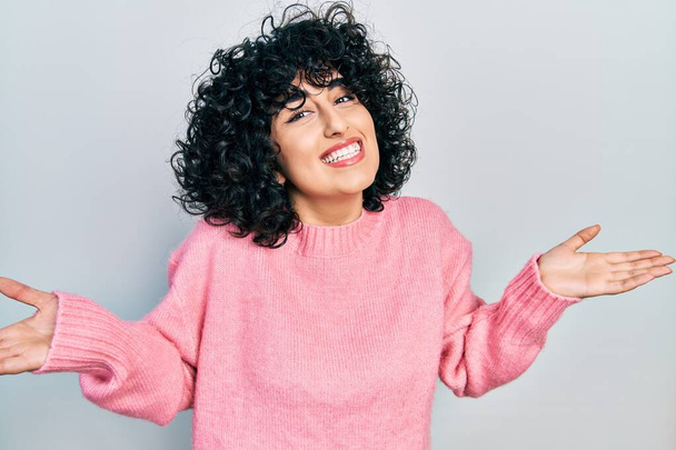Молодая женщина с Ближнего Востока в повседневной одежде улыбается показывая обе руки открытыми ладонями, представляя и сравнивая рекламу и баланс  - Фото, изображение