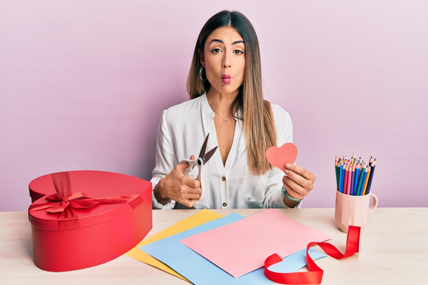 Young hispanic woman making valentine gift sitting on the table making fish face with mouth and squinting eyes, crazy and comical.  - Photo, Image
