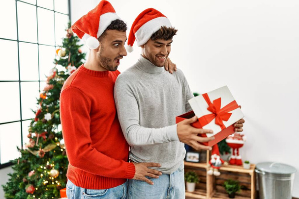 Dva hispánští muži pár překvapení s dárkem stojící u vánočního stromku doma - Fotografie, Obrázek