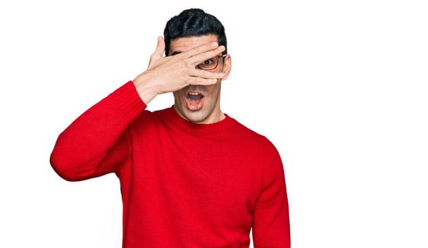 Handsome hispanic man wearing casual clothes and glasses peeking in shock covering face and eyes with hand, looking through fingers with embarrassed expression.  - Photo, Image