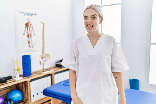 Young caucasian woman working at pain recovery clinic sticking tongue out happy with funny expression. emotion concept.  - Photo, Image