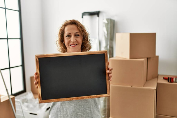 Middle age caucasian woman smiling happy holding empty blackboard at new home. - Photo, Image