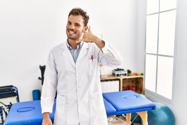 Красивый молодой человек, работающий в клинике болеутоляющих, улыбаясь, делает телефонный жест рукой и пальцами, как будто разговаривает по телефону. коммуникационные концепции.  - Фото, изображение