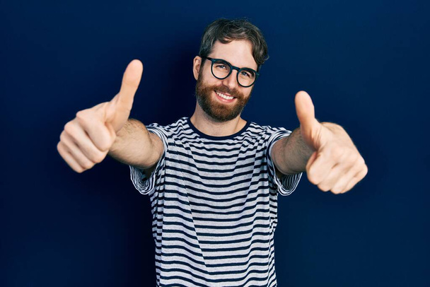 Белый мужчина с бородой в полосатой футболке и очках, одобряющий позитивный жест рукой, улыбается и радуется успеху. жест победителя.  - Фото, изображение