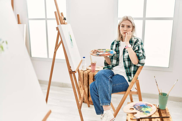 Young artist woman painting on canvas at art studio looking stressed and nervous with hands on mouth biting nails. anxiety problem.  - Fotoğraf, Görsel