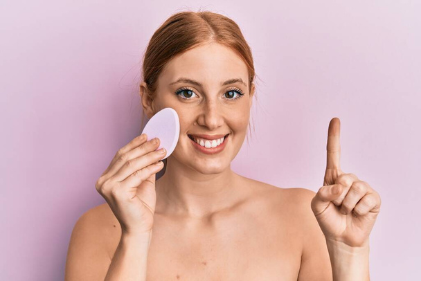 Young irish woman holding makeup sponge smiling with an idea or question pointing finger with happy face, number one  - Photo, Image