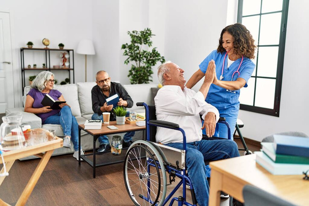Middle age doctor woman and retired man sitting on wheelchair high five at nursing home. - Foto, imagen