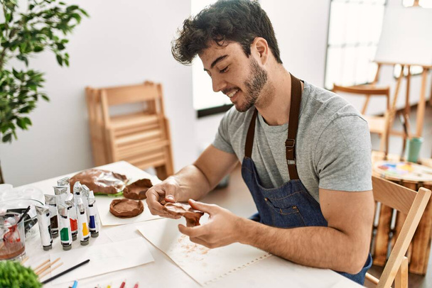 Jovem artista hispânico homem sorrindo feliz modelagem cerâmica no estúdio de arte. - Foto, Imagem
