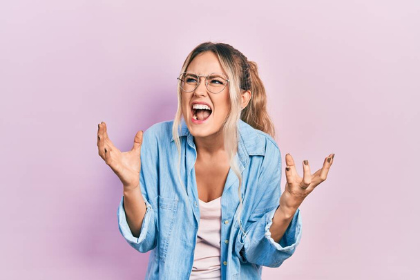 Beautiful young blonde woman wearing casual clothes and glasses crazy and mad shouting and yelling with aggressive expression and arms raised. frustration concept.  - Foto, afbeelding