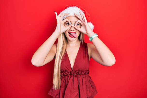 Young blonde girl wearing christmas hat doing ok gesture like binoculars sticking tongue out, eyes looking through fingers. crazy expression.  - Photo, Image