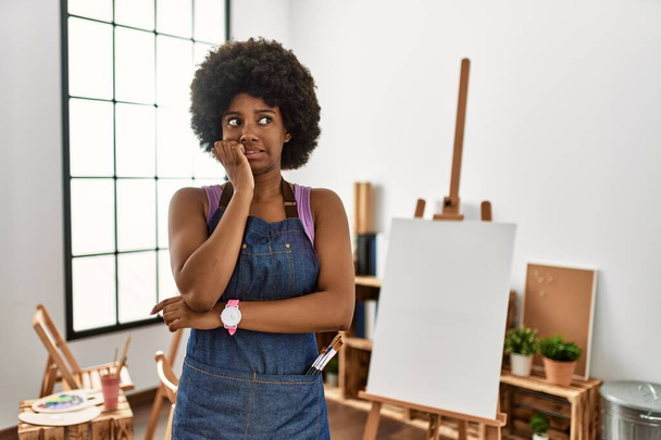 Młoda Afroamerykanka z afro-włosami w studiu sztuki wyglądająca na zestresowaną i zdenerwowaną rękami obgryzającymi paznokcie. niepokój.  - Zdjęcie, obraz