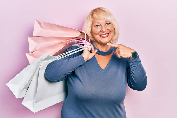 Mujer rubia de mediana edad sosteniendo bolsas de compras sonriendo feliz señalando con la mano y el dedo  - Foto, Imagen