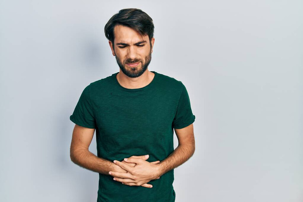 Fiatal spanyol férfi alkalmi fehér pólóban, kézzel a hasán, mert emésztési zavar, fájdalmas betegség rossz közérzet. fájdalom koncepció.  - Fotó, kép