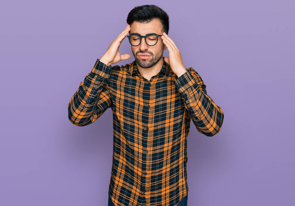 Hombre hispano con barba vistiendo ropa casual y gafas que sufren de dolor de cabeza desesperado y estresado porque el dolor y la migraña. manos en la cabeza.  - Foto, imagen