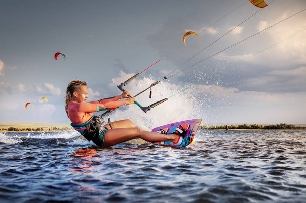 Professional kitesurfer young caucasian woman glides on a board along the sea surface at sunset against the backdrop of beautiful clouds and other kites. Active water sports - Foto, afbeelding