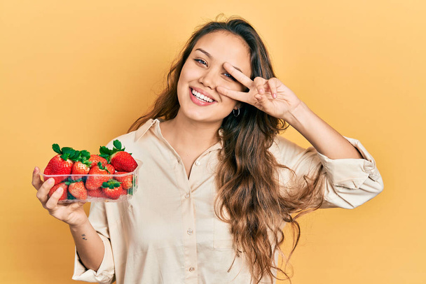 Молодая латиноамериканка, держащая клубнику, делая символ мира с пальцами на лице, улыбаясь радостно показывая победу  - Фото, изображение