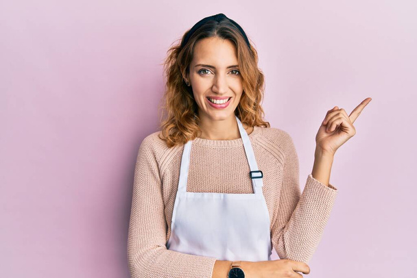 Young caucasian woman wearing apron smiling happy pointing with hand and finger to the side  - Photo, Image