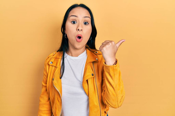 Beautiful hispanic woman with nose piercing wearing yellow leather jacket surprised pointing with hand finger to the side, open mouth amazed expression.  - Photo, Image