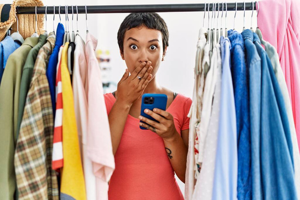 Молода іспаномовна жінка з коротким волоссям шукає одяг на стійці для одягу, використовуючи смартфон, що покриває рот рукою, шокований і боїться помилки. здивований вираз
  - Фото, зображення