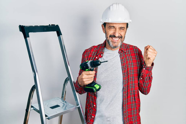 Orta yaşlı İspanyol işçi elinde tornavidayla merdiven kullanıyor gururla bağırıyor, zaferi kutluyor ve başarıyı kollarını kaldırarak kutluyor.  - Fotoğraf, Görsel