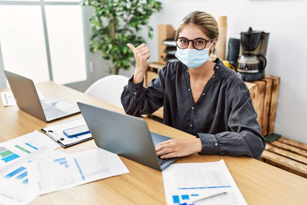 Fiatal szőke nő dolgozik az irodában biztonsági maszkot visel mutatva hüvelykujját az oldalára mosolyogva boldog nyitott szájjal  - Fotó, kép