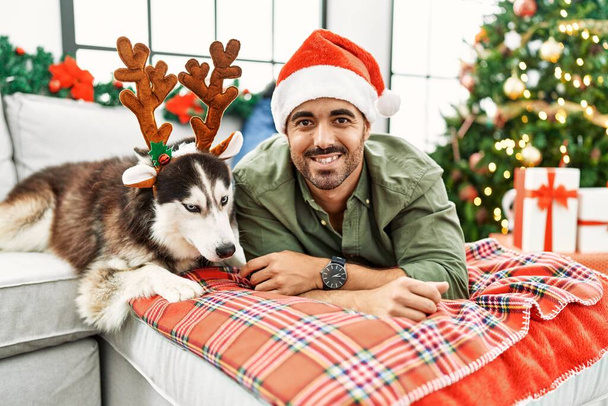 Joven hombre hispano acostado en un sofá con perro con sombrero de reno junto al árbol de Navidad en casa - Foto, Imagen