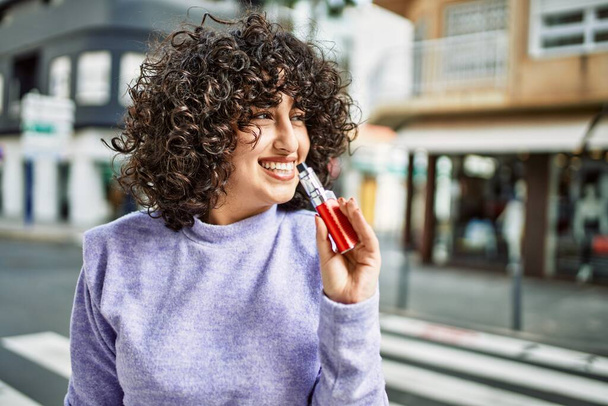 Joven mujer de Oriente Medio sonriendo confiado sosteniendo cigarrillo electrónico en la calle - Foto, Imagen