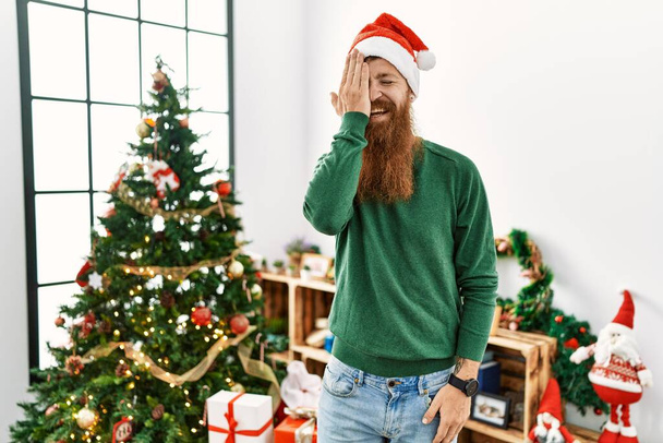 Uomo dai capelli rossi con la barba lunga che indossa il cappello di Natale dall'albero di Natale che copre un occhio con la mano, sorriso fiducioso sul viso ed emozione sorpresa.  - Foto, immagini