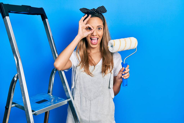 Mooie Latijns-Amerikaanse vrouw door trappen houden roller schilder glimlachend gelukkig doen ok teken met de hand op oog kijken door vingers  - Foto, afbeelding