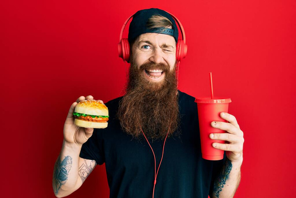 Κοκκινομάλλης με μακριά γενειάδα τρώει ένα νόστιμο κλασικό burger και πίνοντας σόδα να κλείνει το μάτι κοιτάζοντας την κάμερα με σέξι έκφραση, χαρούμενο και χαρούμενο πρόσωπο.  - Φωτογραφία, εικόνα