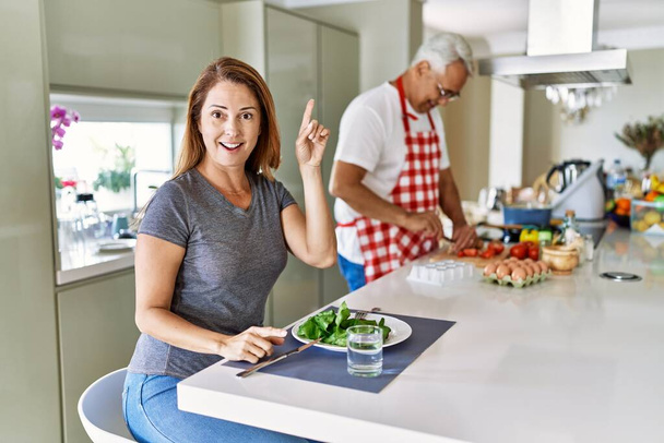 Middle age hispanic couple eating healthy salad at home surprised with an idea or question pointing finger with happy face, number one  - Fotoğraf, Görsel