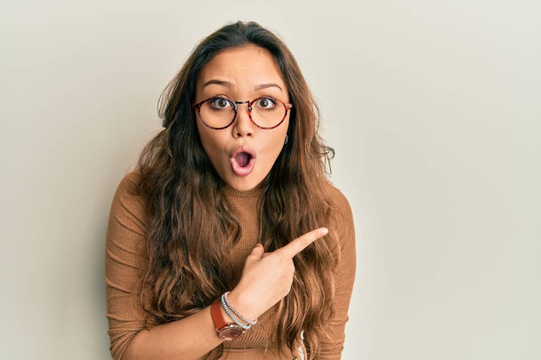 Chica hispana joven con ropa casual y gafas sorprendidas señalando con el dedo hacia un lado, boca abierta expresión asombrada.  - Foto, imagen