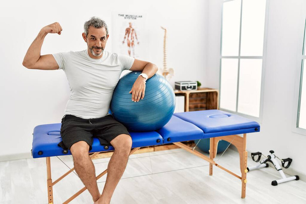 Середнього віку іспаномовний чоловік у клініці відновлення болю тримає пілатес м'яч сильною людиною, показуючи м'язи рук, впевнений і пишається владою
  - Фото, зображення