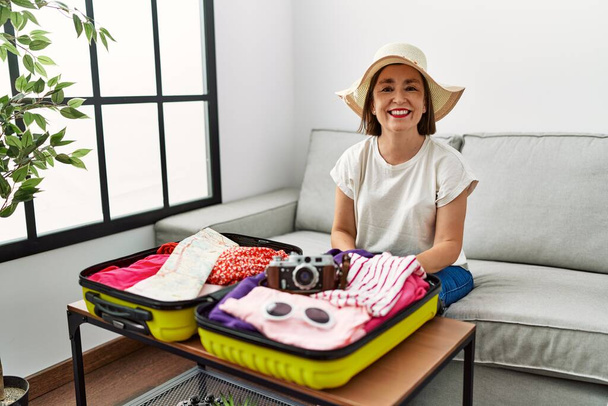 Belle femme hispanique d'âge moyen emballant des vêtements d'été dans une valise avec un sourire heureux et frais sur le visage. chanceux.  - Photo, image