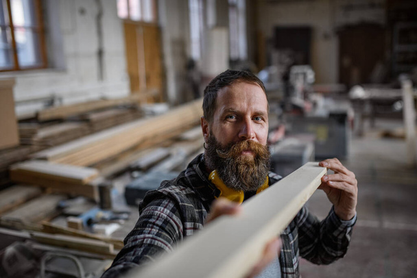 大工のワークショップで木製の板を屋内で運ぶ成熟した男性の大工。中小企業コンセプト. - 写真・画像