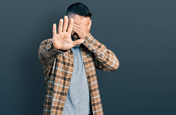 Hombre hispano con barba vistiendo una camisa casual cubriendo los ojos con las manos y haciendo un gesto de stop con expresión triste y de miedo. concepto avergonzado y negativo.  - Foto, Imagen