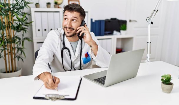 Young hispanic man wearing doctor uniform talking on the smartphone at clinic - Фото, изображение