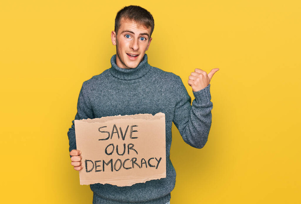 Young blond man holding save our democracy protest banner pointing thumb up to the side smiling happy with open mouth  - Foto, Imagem