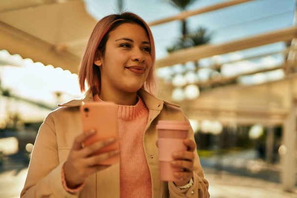 Young hispanic girl using smartphone and drinking coffee at the city. - 写真・画像