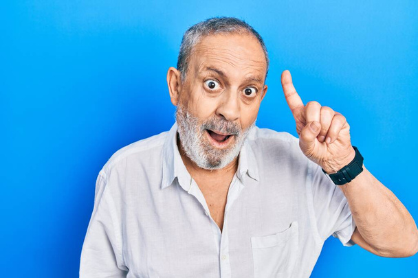 Ein gutaussehender älterer Mann mit Bart trägt ein lässiges weißes Hemd und zeigt mit erhobenem Zeigefinger auf eine erfolgreiche Idee. aufgeregt und glücklich. Nummer eins.  - Foto, Bild