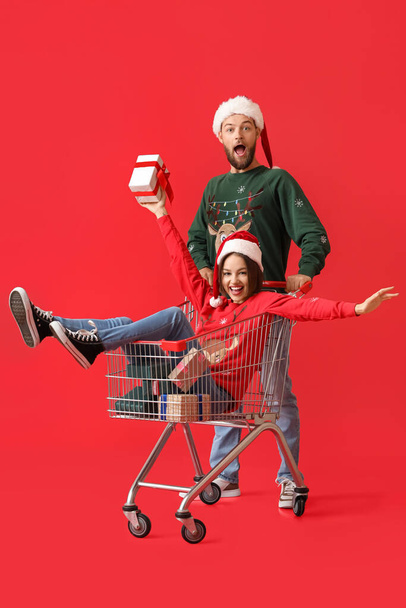 Happy young couple with shopping cart and Christmas gifts on color background - 写真・画像