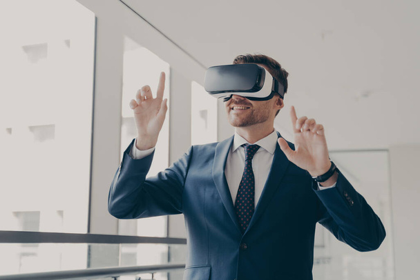Nadšený kancelářský pracovník ve sluchátkách nebo brýlích virtuální reality gestikulující rukama - Fotografie, Obrázek