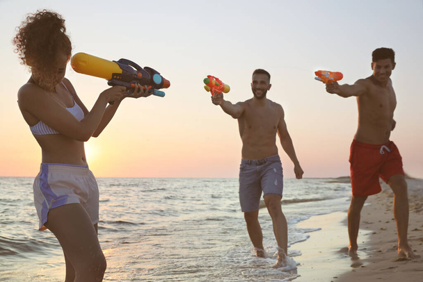 Friends with water guns having fun on beach at sunset - Fotó, kép