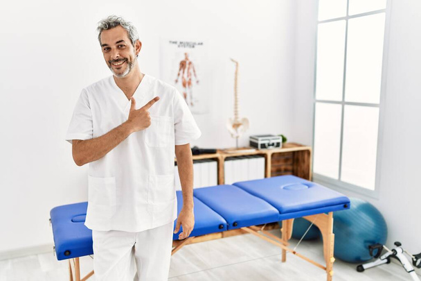 Middle age hispanic therapist man working at pain recovery clinic cheerful with a smile on face pointing with hand and finger up to the side with happy and natural expression  - Photo, Image