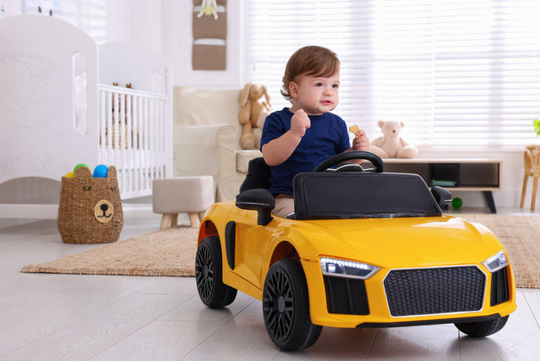 Adorable child with cookie driving yellow toy car in room - Фото, изображение