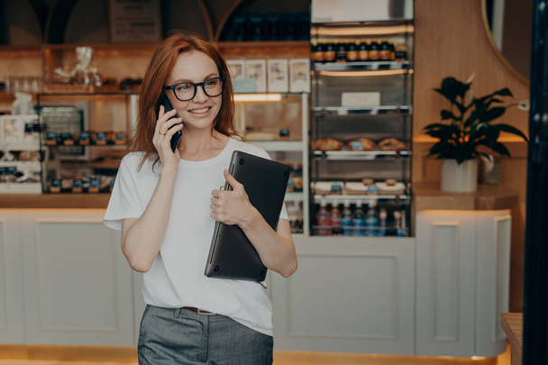Kobieta przedsiębiorca z rudymi włosami ma rozmowę telefoniczną posiada laptopa - Zdjęcie, obraz