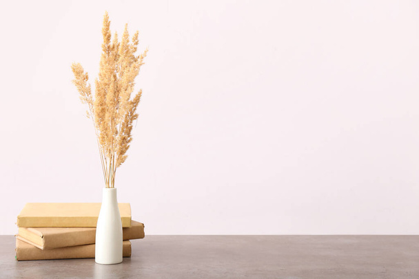 Vase with beautiful dried spikelets and books on table near light wall - 写真・画像