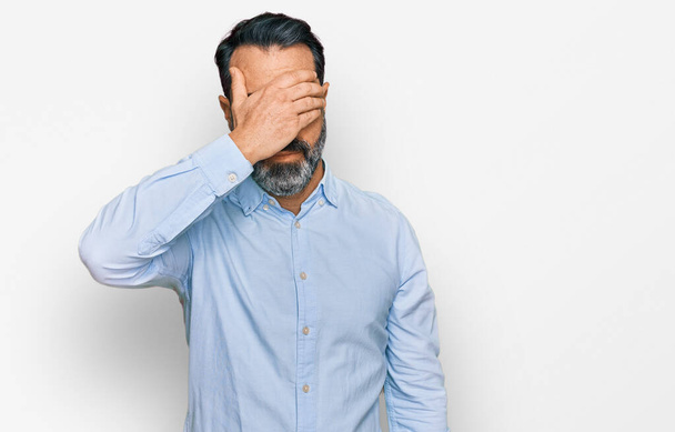 Middle aged man with beard wearing business shirt covering eyes with hand, looking serious and sad. sightless, hiding and rejection concept  - Photo, Image
