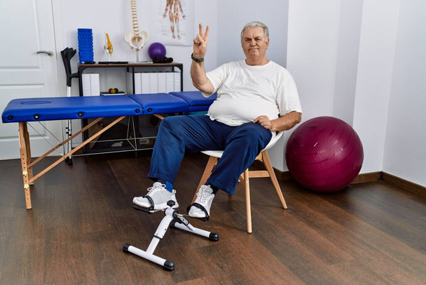 Uomo anziano caucasico presso la clinica di fisioterapia utilizzando l'esercizio a pedali mostrando e indicando con le dita numero due sorridendo fiducioso e felice.  - Foto, immagini
